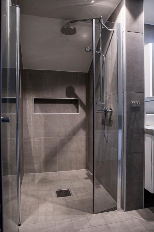 A walk-in shower with glass doors, grey wall tiles and a recessed shelf