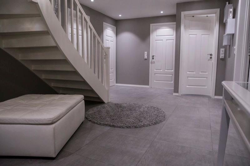 A hallway with large grey floor tiles, a white staircase and white doors